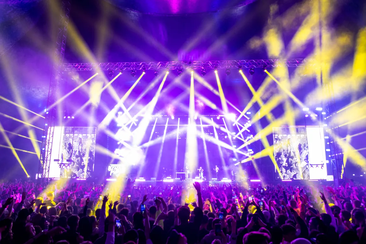 Festival crowd with raised hands facing a stage lit by purple and yellow light beams at night